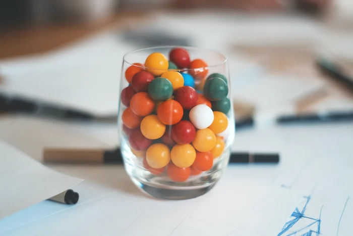 Drinking glass containing colourful bubble gum balls sat atop a desk with scattered papers